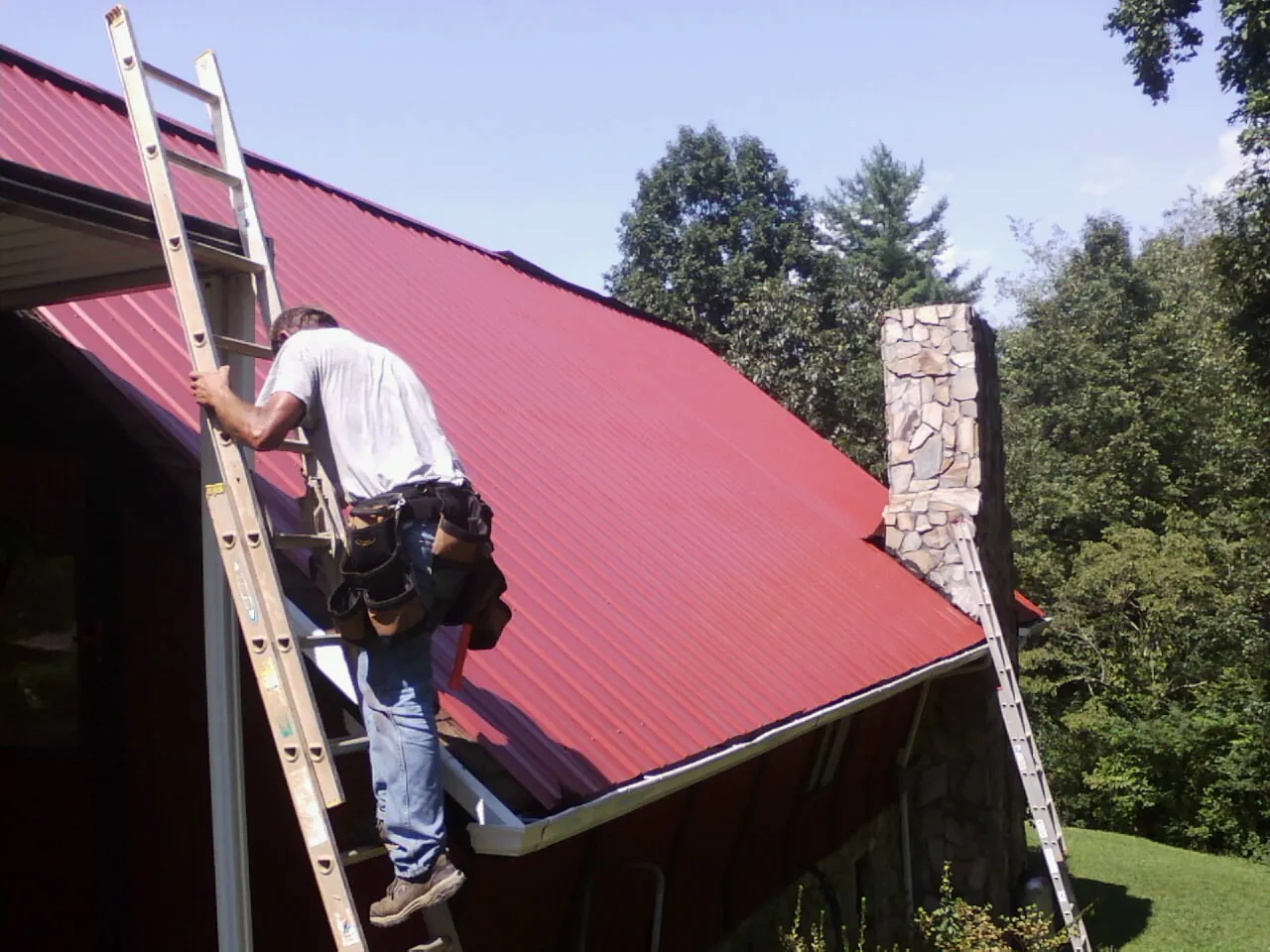 Roofer on a ladder beside a red metal roof and stone chimney — Soffit & Fascia Repair by Roofing Brothers in Wellsville